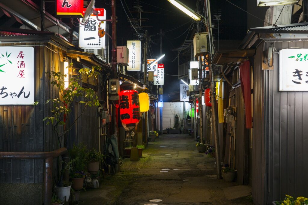 A narrow alley of izakaya and sushi bars glowing with signs and lanterns at night