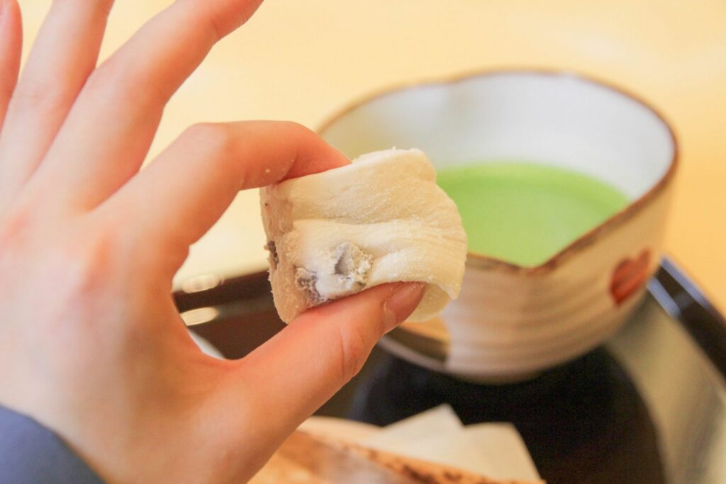 A hand holding a soft white yumochi mochi with a bowl of green matcha behind