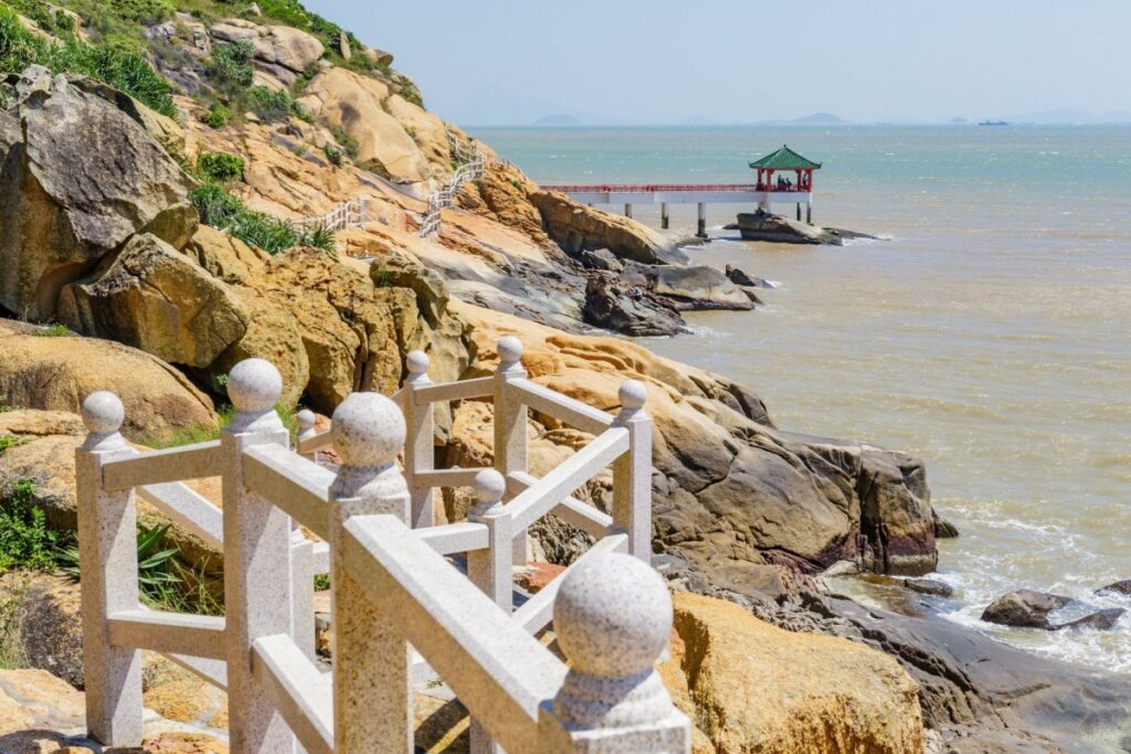 A Chinese pavilion on a rocky pier along Macau's Coloane island coastline under a hazy sky