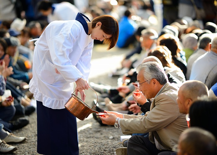 A shrine attendant pouring doburoku sake from a copper pot for seated visitors at the festival