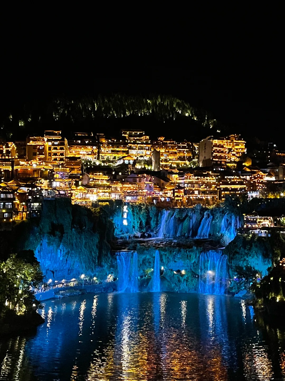 Furong Ancient Town lit gold on the cliff above its blue-lit waterfall at night
