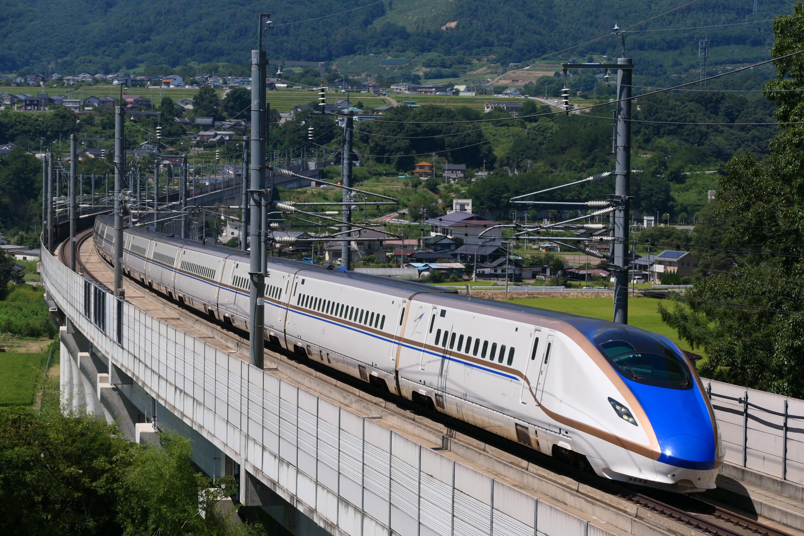 A white and blue Hokuriku Shinkansen train crossing a viaduct through green countryside