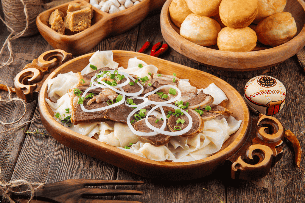 Sliced boiled meat and onions over wide flat noodles on a wooden platter