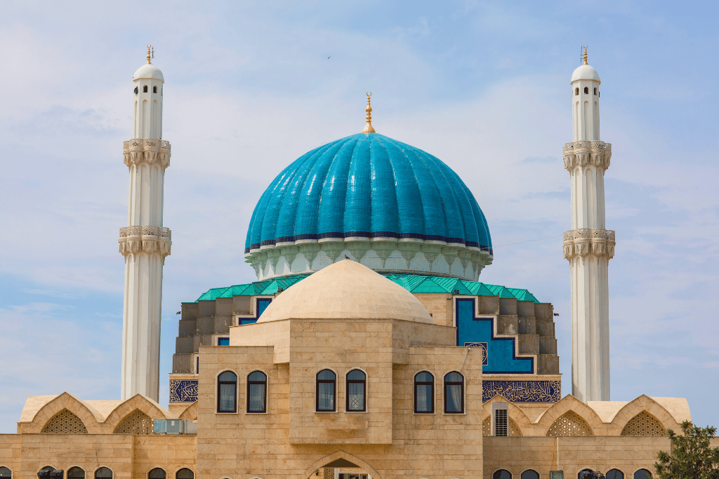 A sandstone mosque with a ribbed turquoise dome and two white minarets under a blue sky
