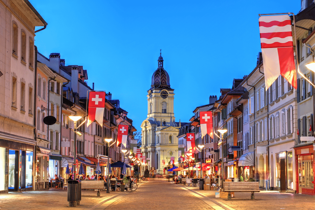 A cobbled old-town street strung with Swiss flags at dusk, a domed church tower ahead