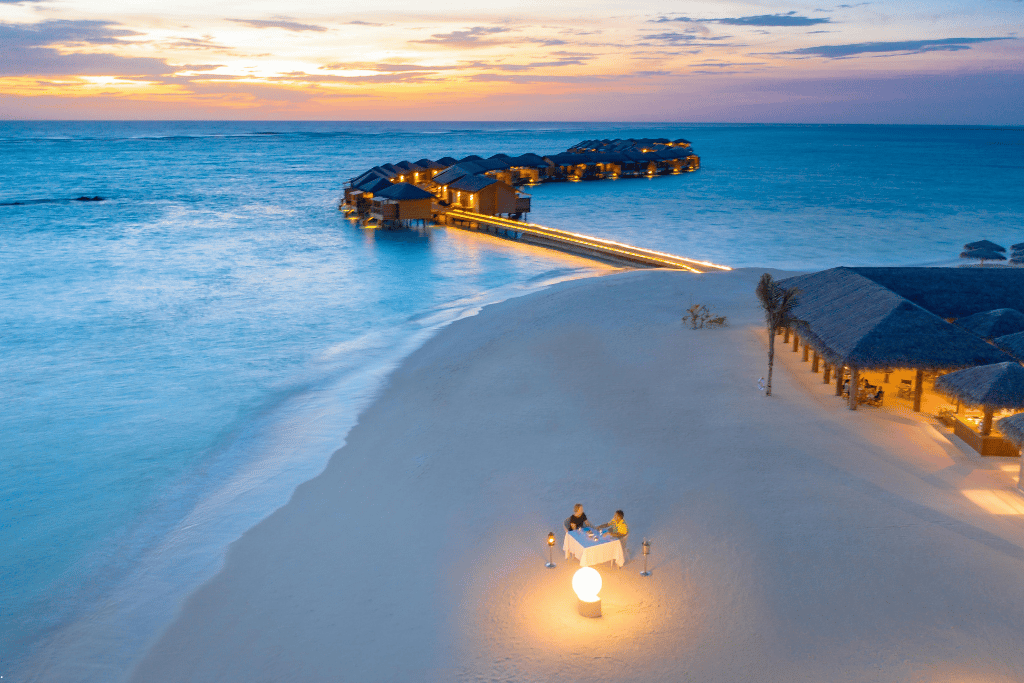 A couple at a candlelit dinner on a white-sand beach at dusk, lit overwater villas along a jetty behind