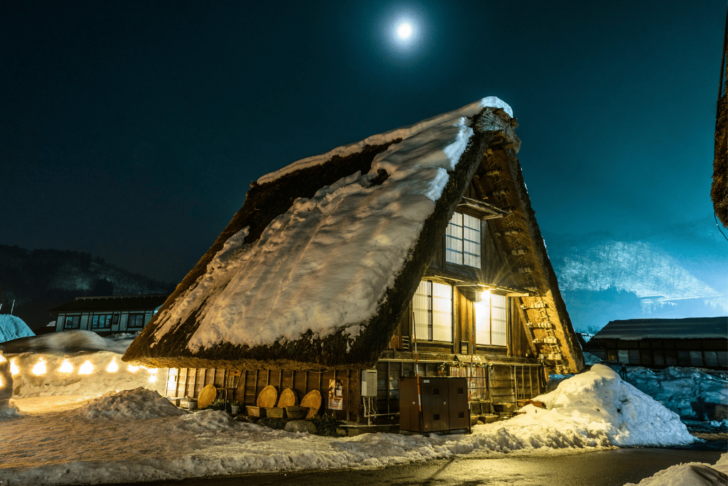 A snow-covered gassho-zukuri farmhouse lit up at night under a full moon