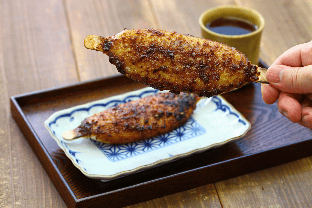 A grilled gohei mochi rice skewer glazed with miso, held over a plate with dipping sauce