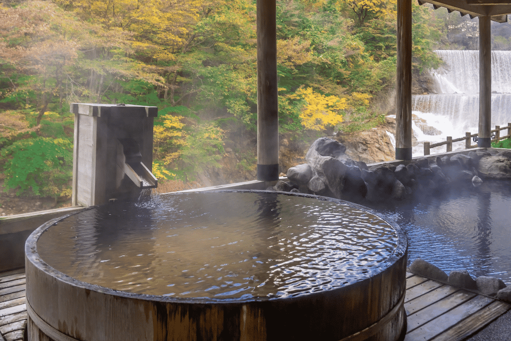 A steaming round wooden open-air hot-spring bath with autumn trees and a waterfall behind
