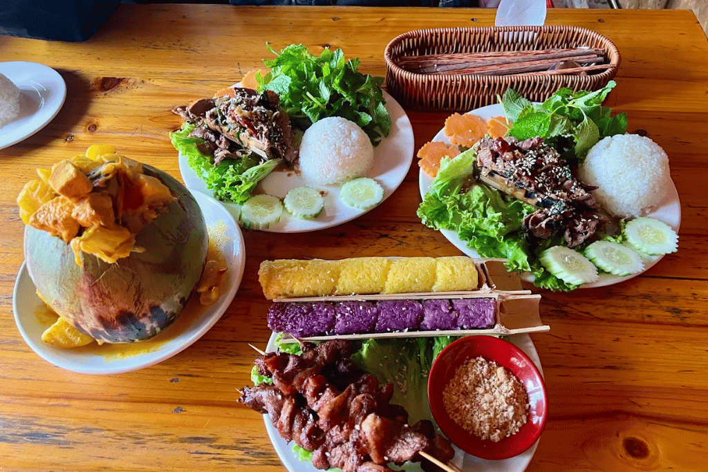 A wooden table spread of chicken curry in a coconut, grilled meats with rice, and colourful bamboo-tube sticky rice