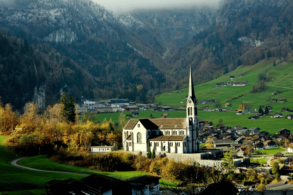 A white church spire above a green valley village, early snow dusting the peaks behind