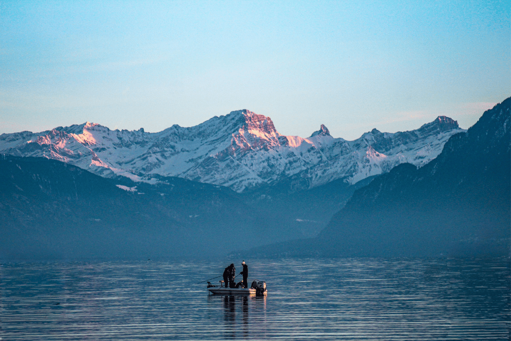 Two people in a small boat on a calm lake at dusk, snow-capped peaks glowing behind