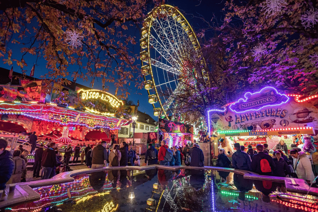 A night funfair with a lit Ferris wheel and glowing ride stalls under autumn trees