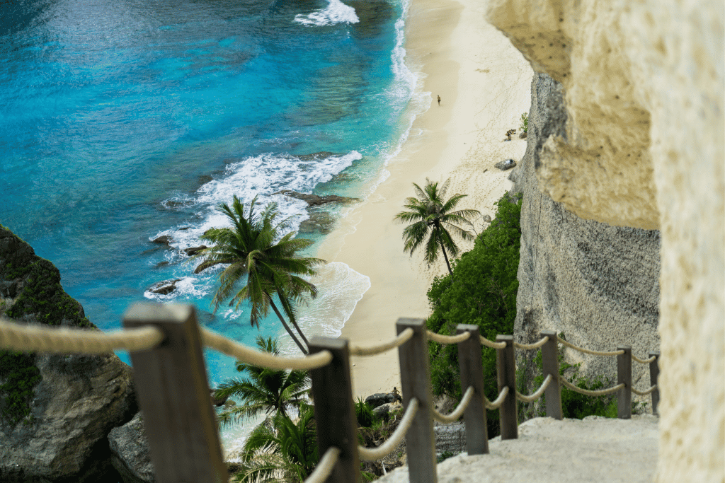 View down a rope-railed cliff stairway to a palm-fringed white-sand beach and turquoise sea