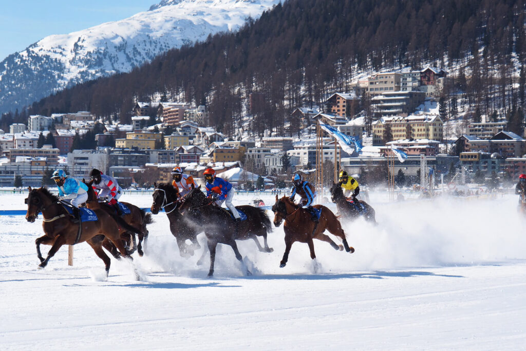 Jockeys racing horses across the snow at White Turf, with St Moritz behind