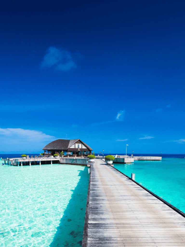A long wooden jetty leading to a thatched overwater restaurant over a bright turquoise lagoon under blue sky