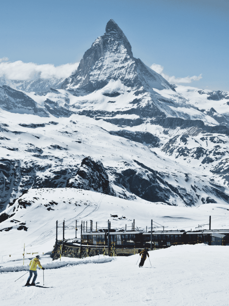 Two skiers on a snowy piste below the Matterhorn, with a mountain railway train