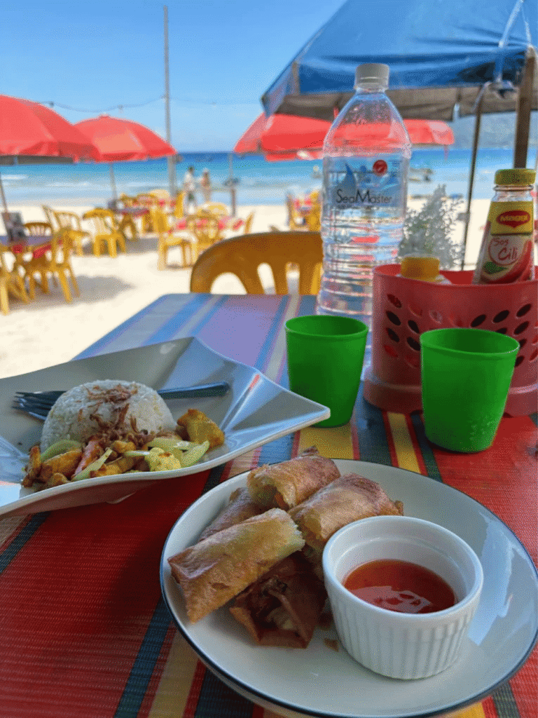 A plate of rice and spring rolls with chilli sauce on a beachside table under red umbrellas at Long Beach