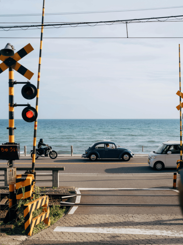 A vintage car and a motorcycle waiting at a seaside Enoden railway crossing near Kamakura