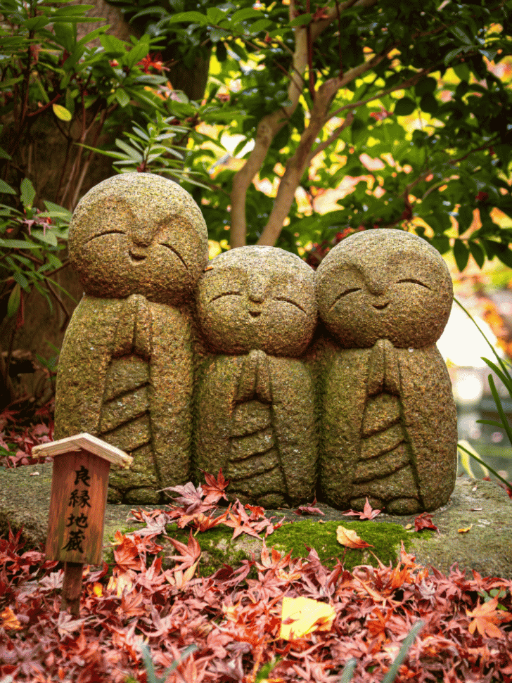 Three smiling stone Jizo statues among red autumn maple leaves at a Kamakura temple