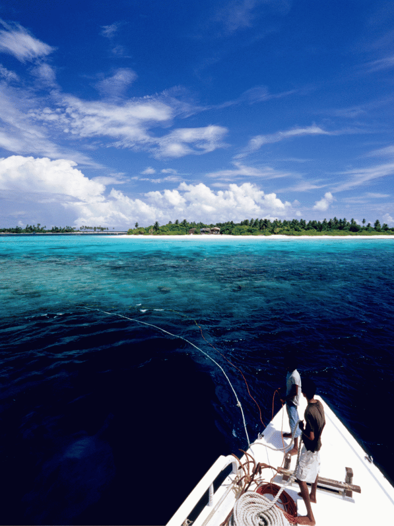 View from a boat's bow of two crew approaching a palm-fringed Maldives island across turquoise reef