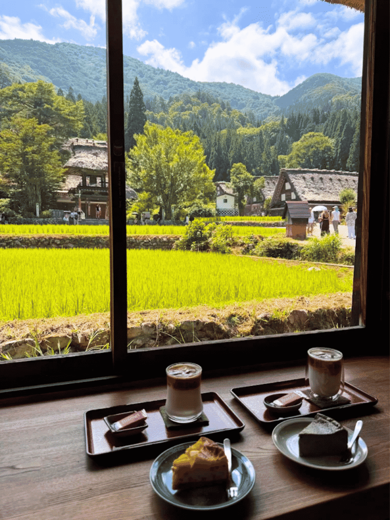 Coffee and cake by a gassho-house cafe window overlooking green rice paddies and thatched roofs