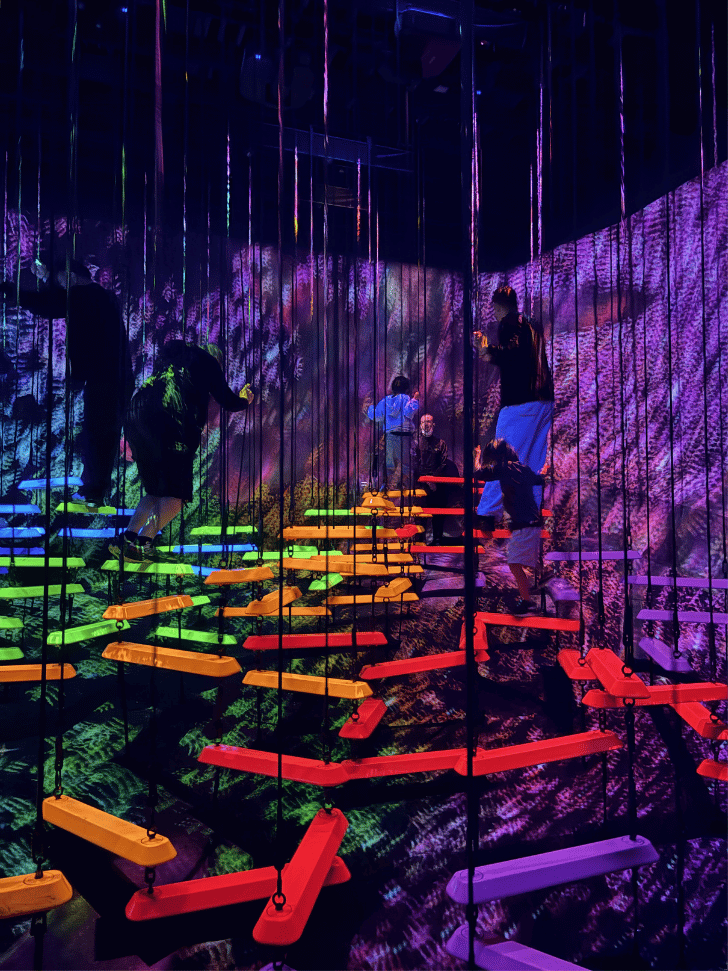 Blue flowing light lines swirling across the floor of a dark teamLab room with visitors