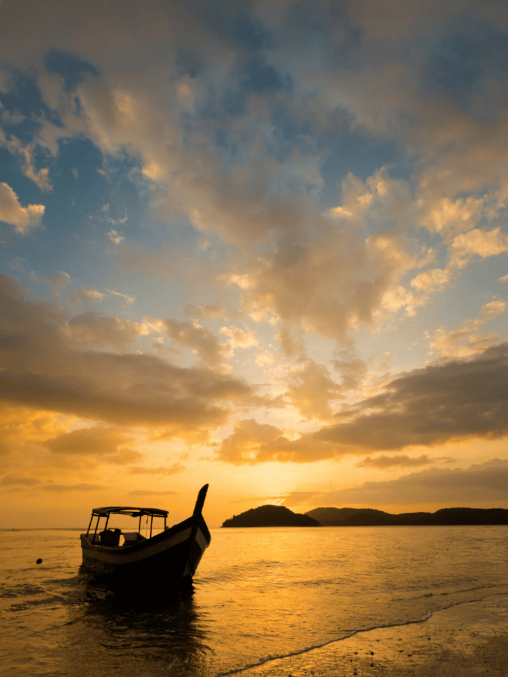 Wooden boat silhouetted against a golden sunset over the sea at Pantai Cenang