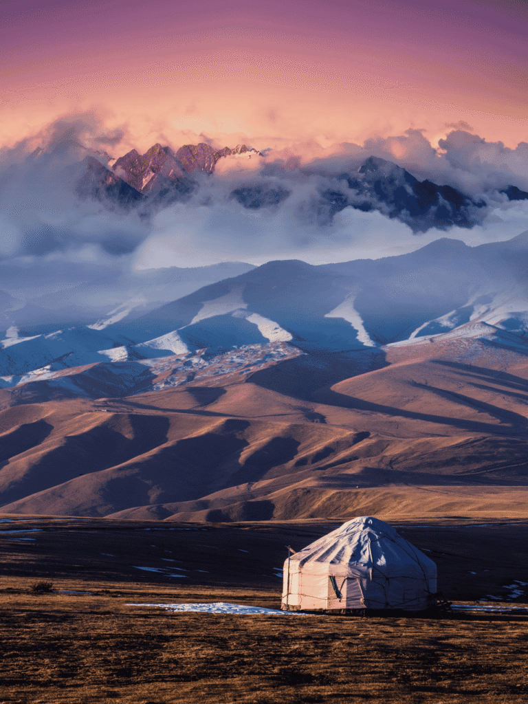 A lone yurt on the steppe below cloud-wrapped mountains at pink dawn