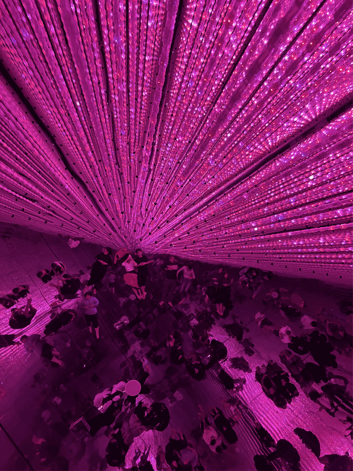 Pink radiating light strands fanning above a crowd reflected on a mirrored floor at teamLab Planets