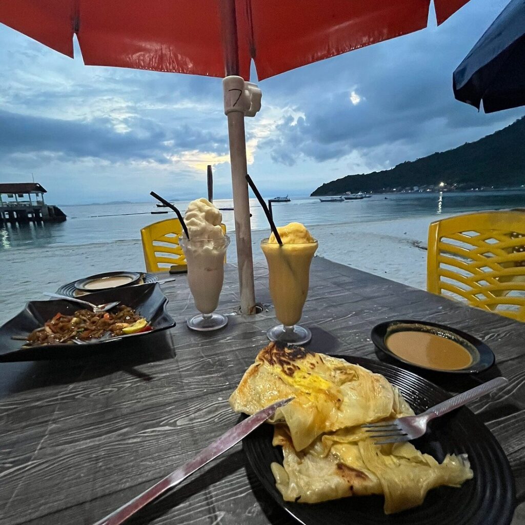 Two fruit shakes, roti canai and a noodle dish on a beachside table at dusk on Perhentian Besar