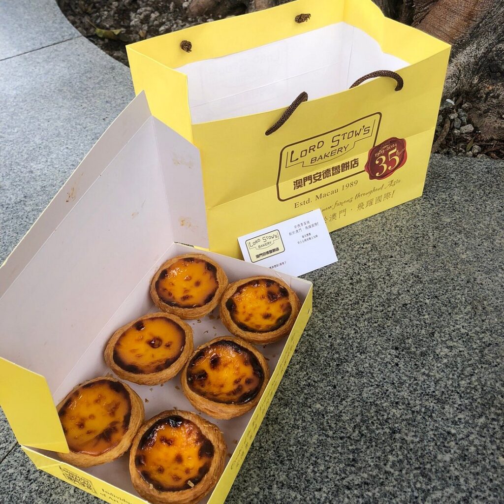 A box of six Portuguese egg tarts beside a yellow Lord Stow's Bakery bag in Macau