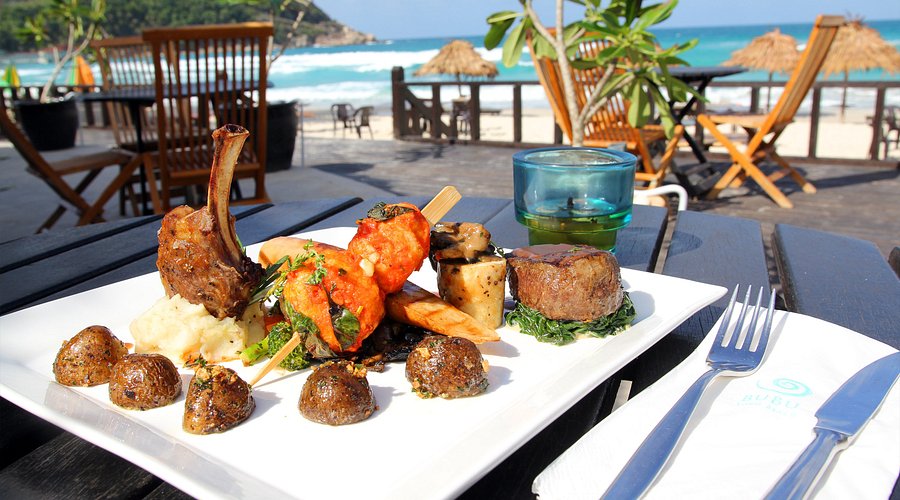 A grilled meat platter on a white plate at a beachfront table overlooking Long Beach