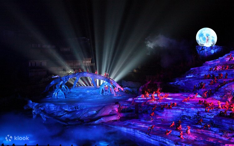 Open-air night stage show lit blue and purple below Tianmen Mountain with a moon prop