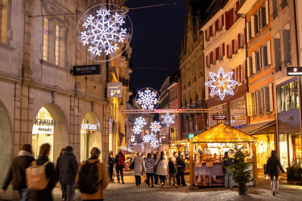 An old-town street hung with snowflake lights, a wooden market chalet and shoppers at night
