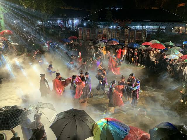 Ethnic-minority performers dancing in the mist under coloured lights as an umbrella-holding crowd watches