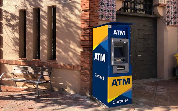 A blue and yellow Euronet ATM standing on a pavement beside a bench