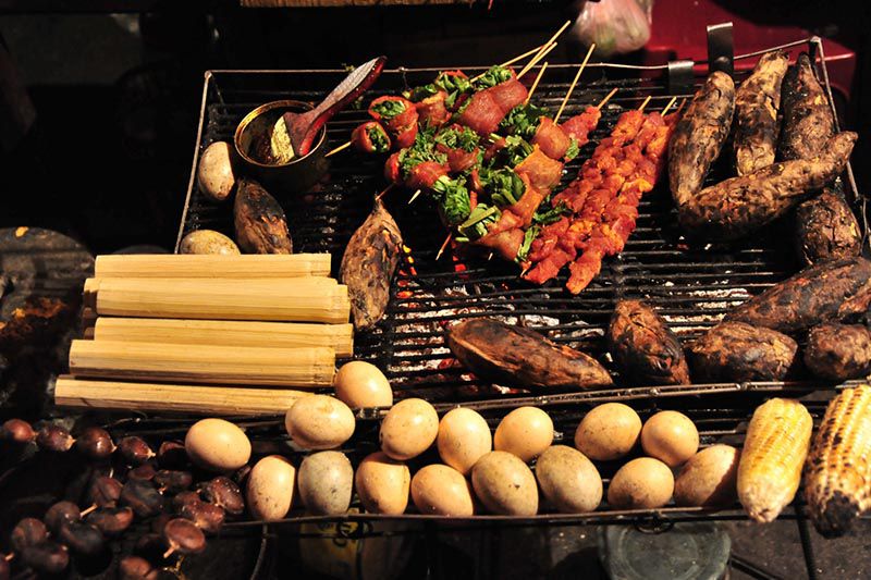 A charcoal grill loaded with marinated meat skewers, grilled eggs, corn and bamboo-rice tubes at a night market