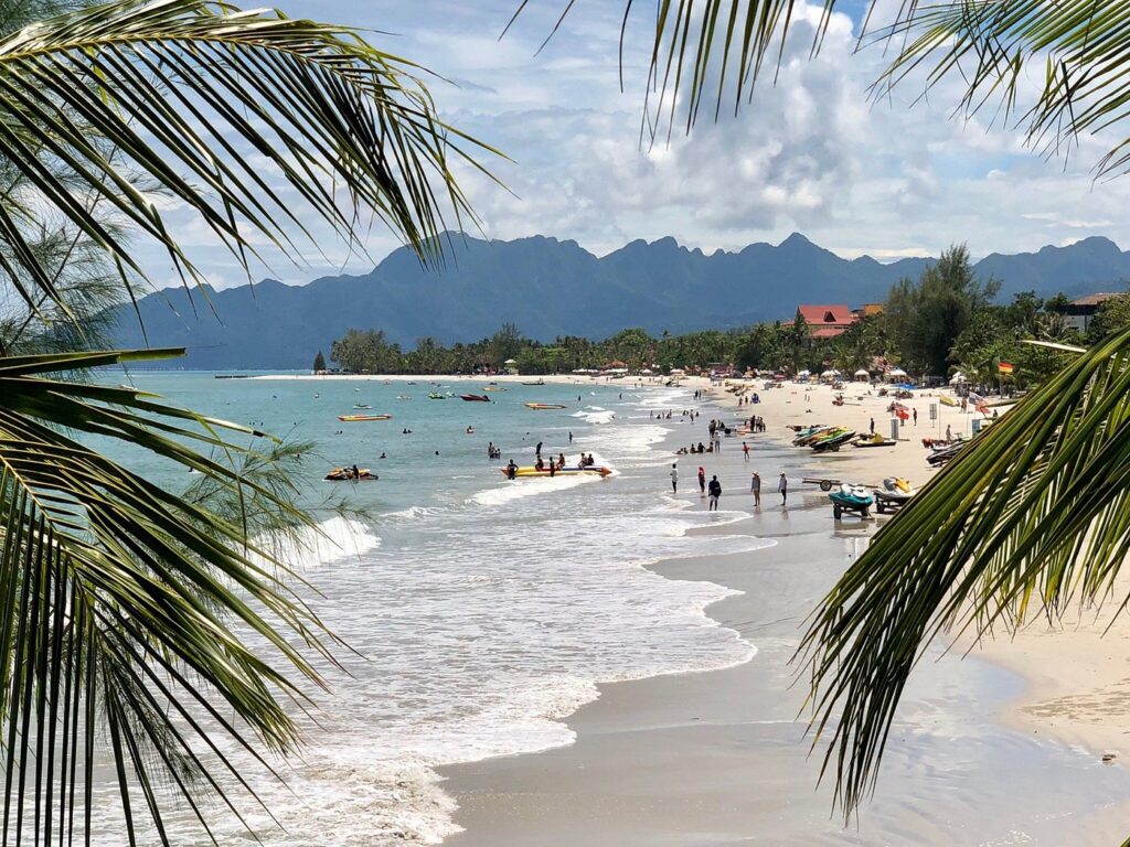 Busy Pantai Cenang beach with water sports and mountains behind, framed by palm fronds