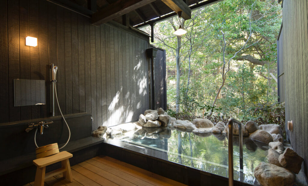A stone open-air hot-spring bath beside a wooden deck, looking onto green forest