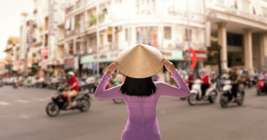 Woman in a purple ao dai and conical hat facing a busy motorbike-filled street