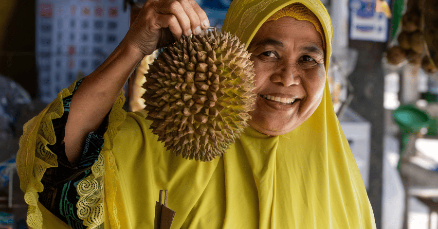 A smiling durian seller in a yellow headscarf holds up a whole durian at her market stall