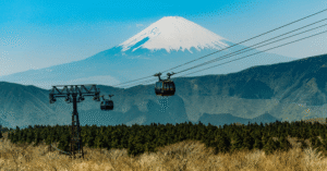 Snow-capped Mt Fuji rising behind two Hakone Ropeway gondolas over forested hills