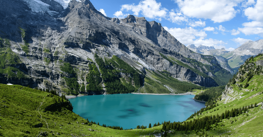 A turquoise alpine lake ringed by craggy grey peaks and green mountain meadows
