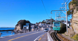 A green Enoden train on the coastal road at Kamakura, with the sea and a small headland behind