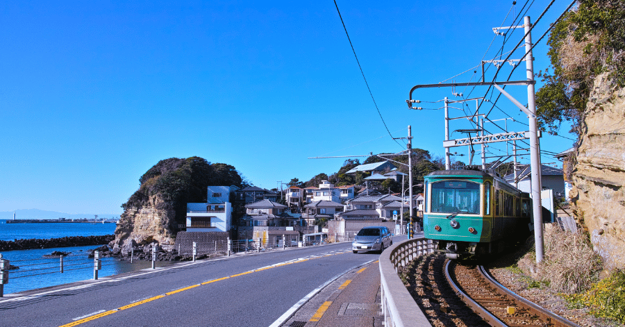 A green Enoden train on the coastal road at Kamakura, with the sea and a small headland behind