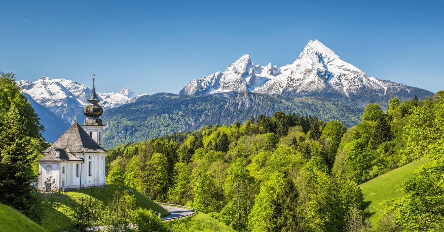 White onion-domed church on a green hillside below the snow-capped Watzmann in Bavaria