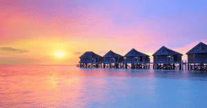 A row of thatched overwater villas on stilts at sunset, pastel pink and orange sky over calm water