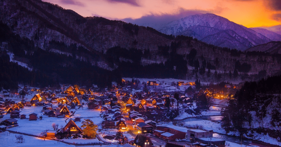 Snow-covered Shirakawago village at dusk with lit gassho-zukuri farmhouses below glowing mountains