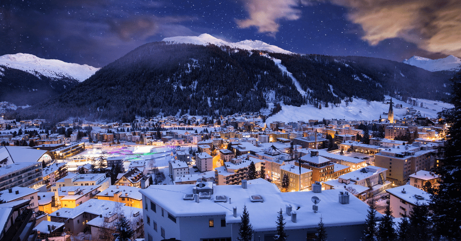 A snow-covered Swiss mountain town glowing at dusk beneath forested ski slopes